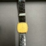 オメガ デ・ヴィルのレディース腕時計。ゴールドケースにブラックレザーベルトの上品なモデルが撮影台に置かれている写真。