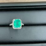 黒いジュエリートレーの上に置かれたエメラルドのダイヤモンドリング。中央に四角形のエメラルド、その周囲を小粒ダイヤが取り巻くデザイン。金属部分は明るい白色でK18またはプラチナ台と思われる。